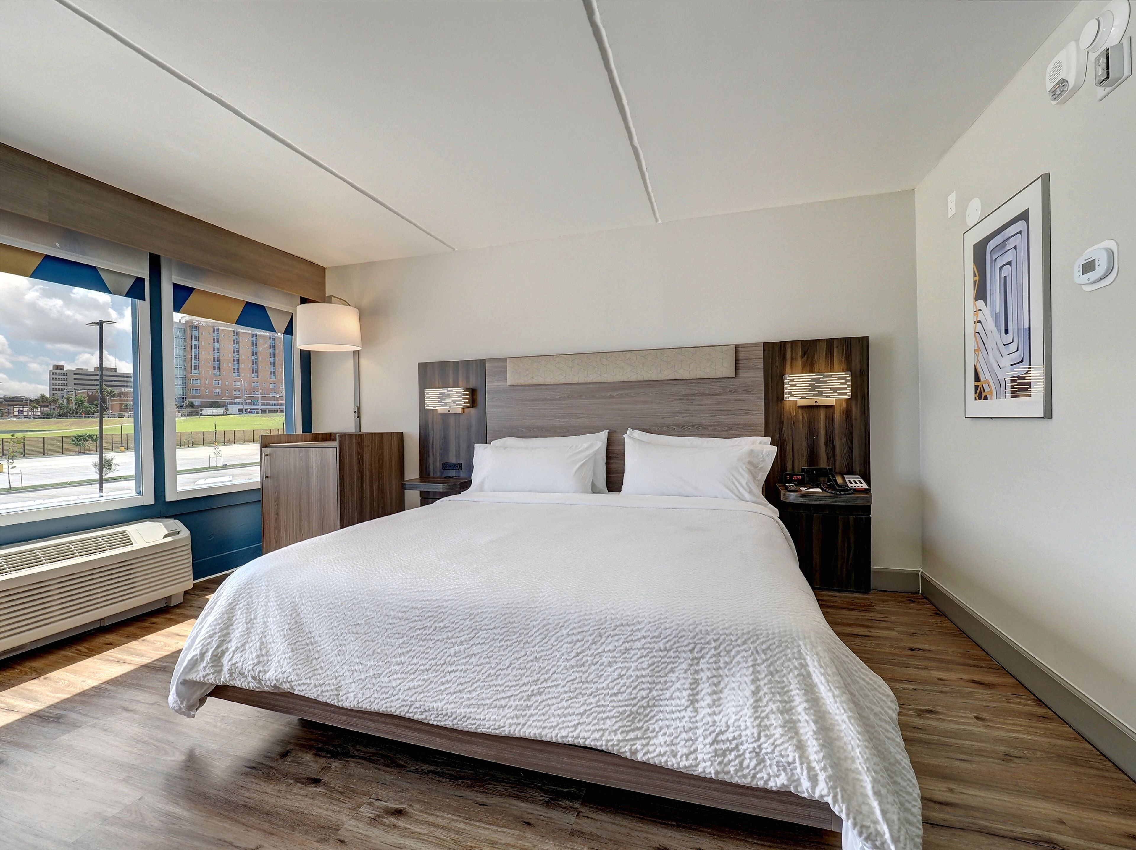 Una habitación de hotel moderna con una cama grande, un escritorio y una ventana con vistas a un edificio y un estacionamiento.