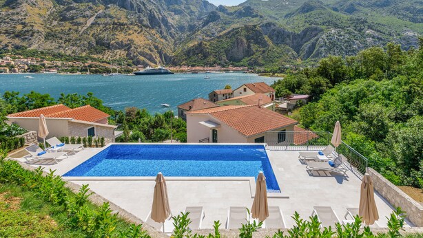 A swimming pool with lounge chairs and umbrellas, surrounded by greenery and overlooking a body of water with boats and buildings in the distance. - Apartments Doncic (Kotor)