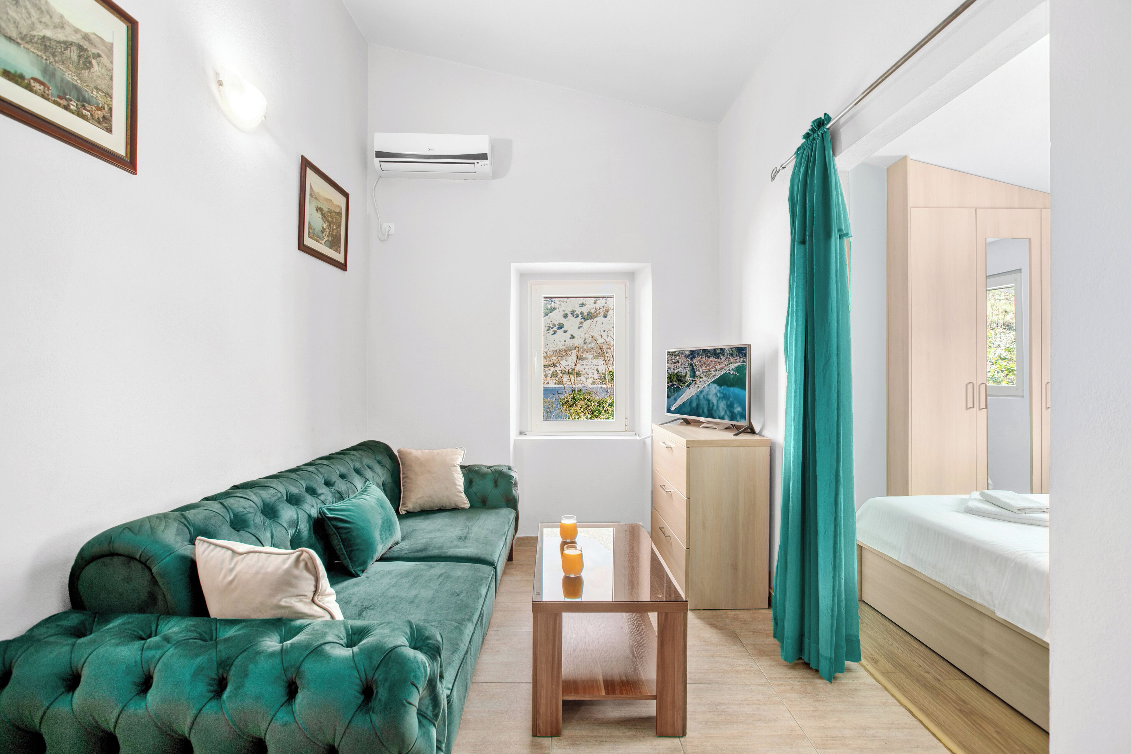 A modern room with a green sofa, a wooden table with a glass of orange juice, a bed with a teal curtain, and a TV on a wooden cabinet.