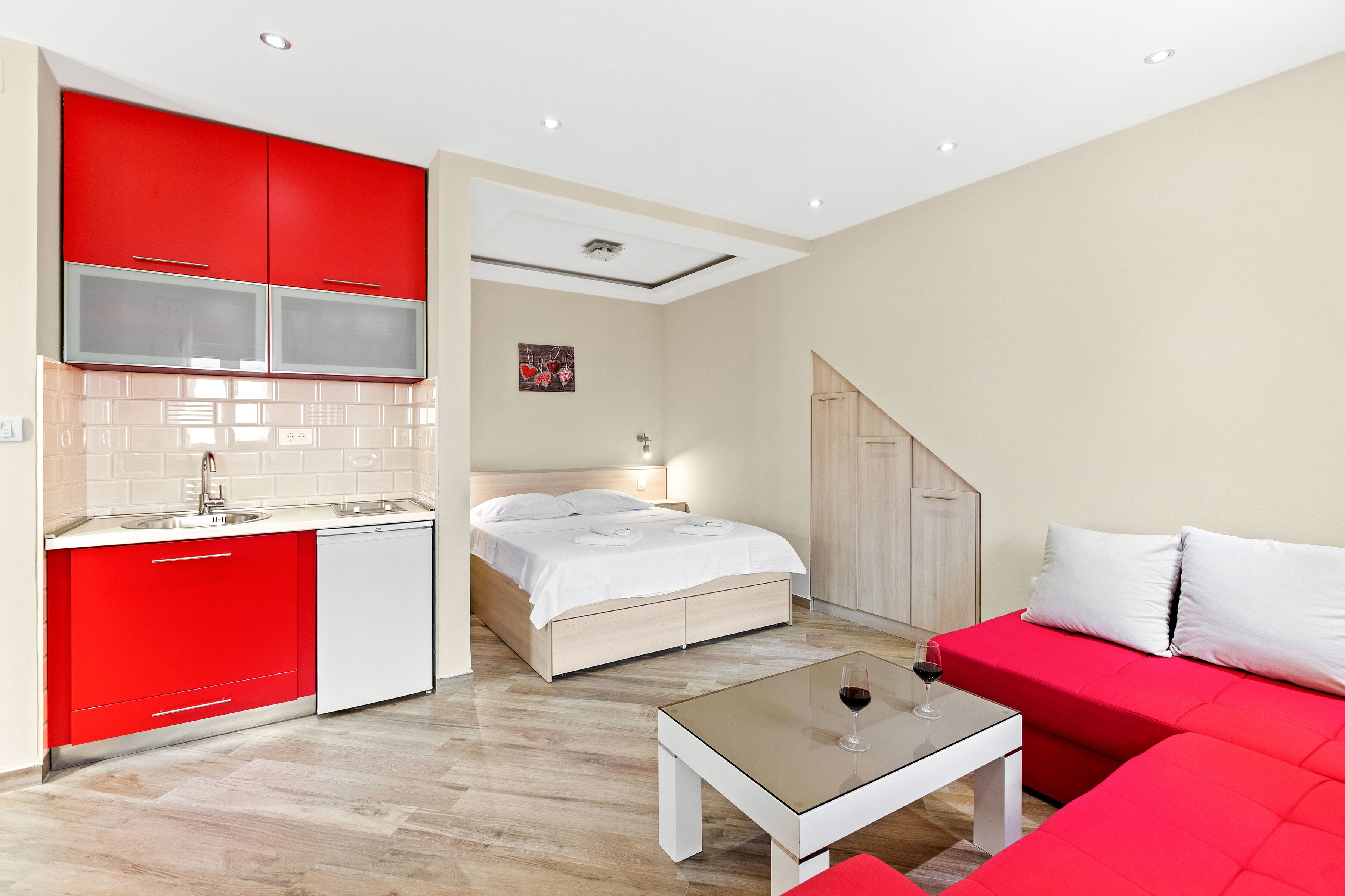 A compact hotel room with a kitchenette, a bed, a sofa, and a small table with a glass of wine.