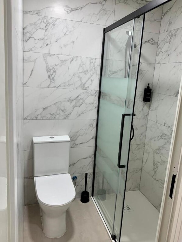 A bathroom with a toilet, a glass-enclosed shower, and marble walls. - Boga Hostel (Madrid)