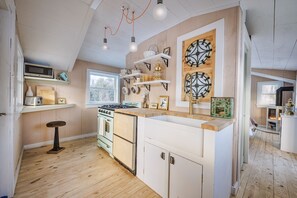 Een keuken met houten vloeren, witte kastjes, een fornuis en een magnetron. Er zijn planken met servies en een klok aan de muur.