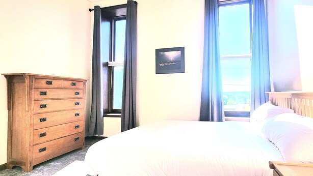 Een slaapkamer met een houten dressoir, een bed met wit beddengoed, een ingelijste foto aan de muur en een raam met gordijnen.