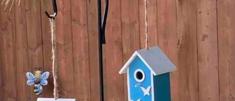 A birdhouse and feeder hanging from a metal hook against a wooden fence.