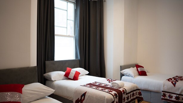 A hotel room with two beds, each with a red and white pillow, a quilt, and a window with curtains. - Carlton Residence Sleeps 6 (London)