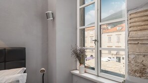 Una esquina de habitación con una cama, una mesita, un florero con flores y una ventana que ofrece vistas a edificios.