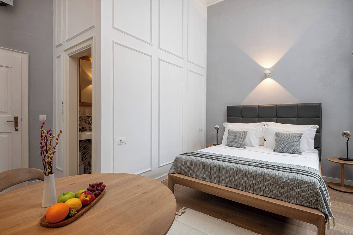 A bedroom with a bed, bedside table, and a nightstand. There is a dining table with fruit and a vase in the foreground.