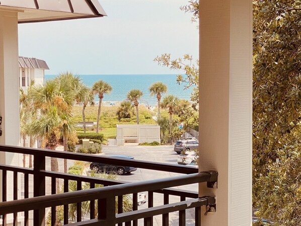 A balcony with a view of the ocean, palm trees, and a parking area.