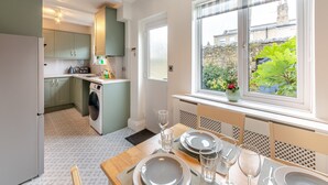 A compact kitchen with a washing machine, a dining area with a table set for two, and a window with a view of greenery and buildings. - 3 bedroom accommodation in Alnwick (Alnwick)