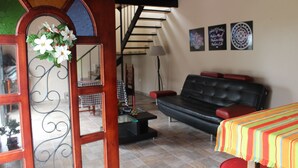 A living room with a black leather sofa, a wooden coffee table, and a colorful striped tablecloth. There is a staircase in the background leading to an upper level. - Bhakti Coffee Farm (Zipacon)