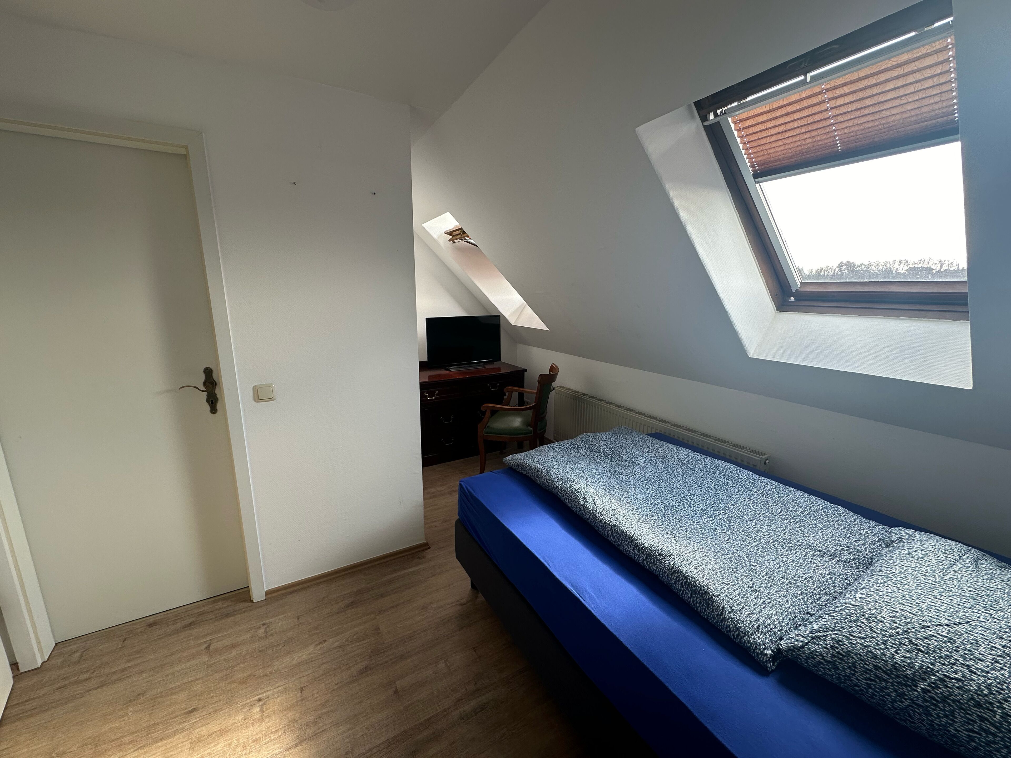 A small, well-lit room with a sloped ceiling, a single bed with a blue mattress, a desk with a chair, and a television.