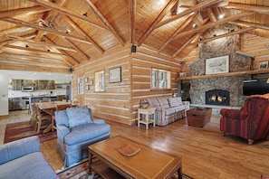 A log cabin interior with a stone fireplace, wooden furniture, and a kitchen area. - Historical Crocker Ranch - Rowe Cabin - 23-ZONE3423 (Estes Park)