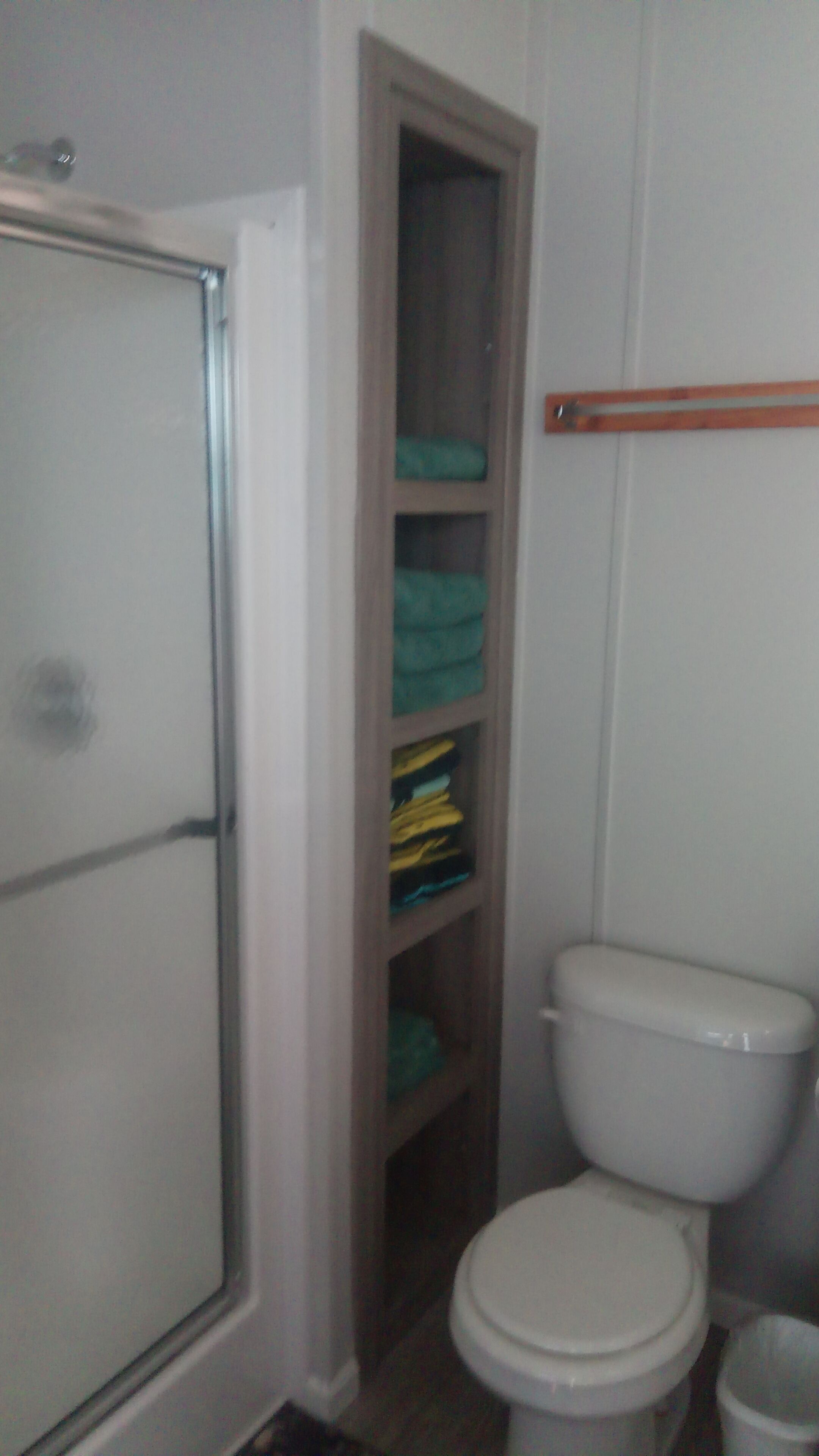 Un baño con inodoro blanco, cabina de ducha y estante empotrado con toallas dobladas.