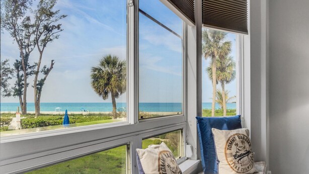 A room with large windows overlooking a beach, featuring a view of the ocean and palm trees. There are two pillows on the windowsill, one with a beach-themed design.