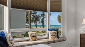 A bedroom with a large window overlooking a beach, a bed with a white comforter, and two pillows with a beach-themed design.
