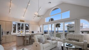 A spacious living area with a large window overlooking the ocean, a ceiling fan, and a modern kitchen with bar stools.