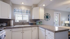 A kitchen with white cabinets, a granite countertop, a built-in dishwasher, a window with blinds, a decorative compass wall art, and a small island with a sink. - 2 Bedroom End Unit, Hot Tub, Parking, - Steps from the Beach! (Holmes Beach)