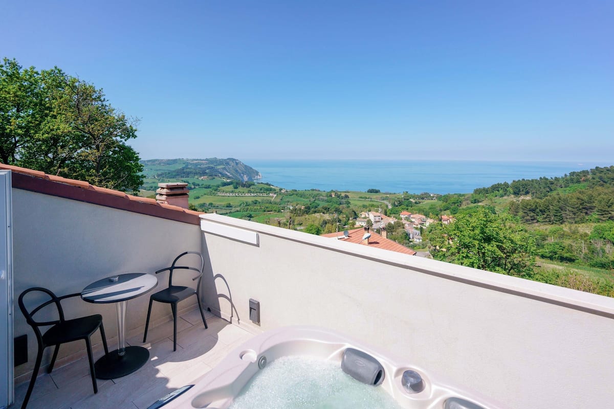 A balcony with a hot tub, two chairs, and a view of the sea and greenery.