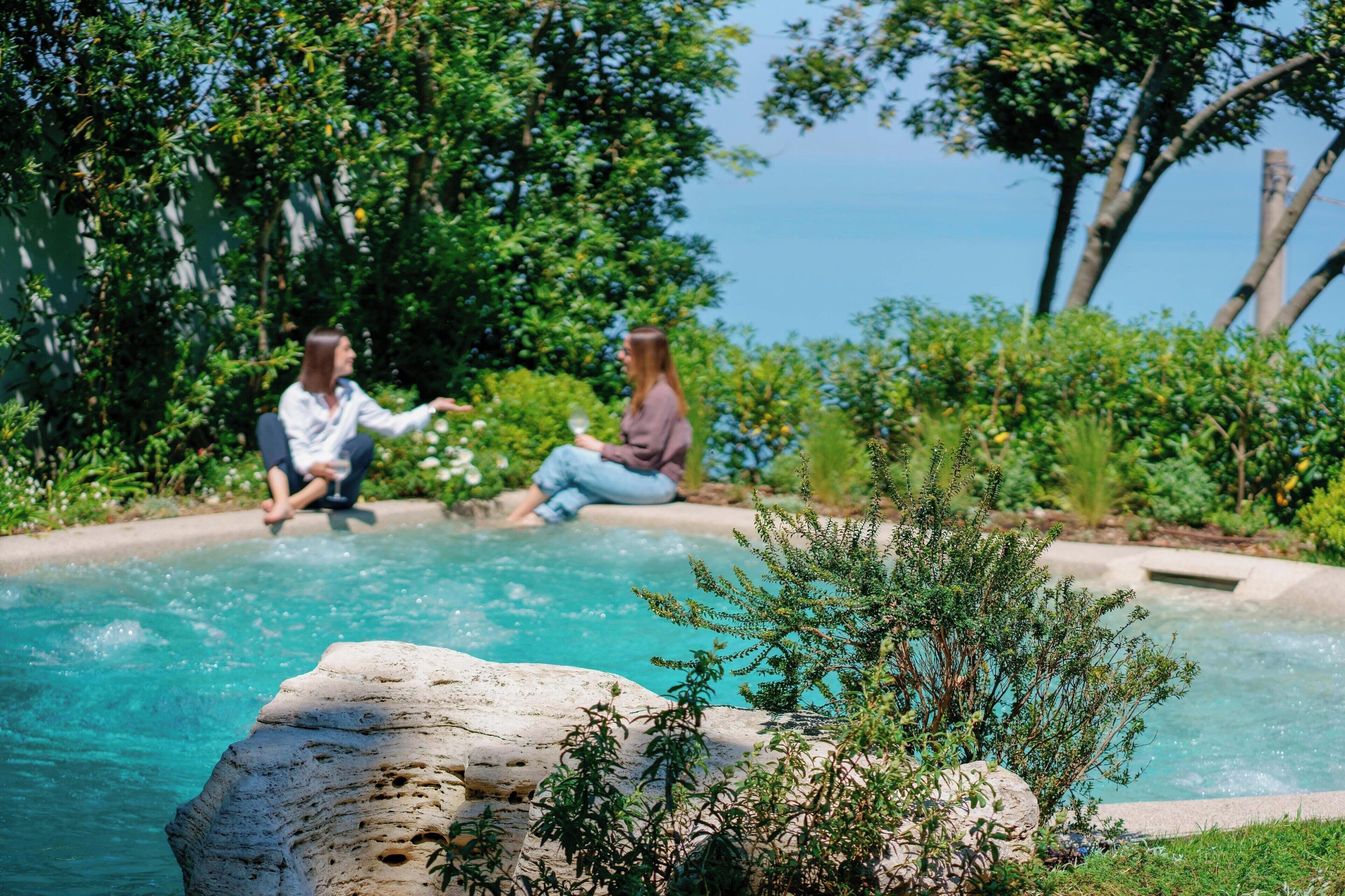 A pool with two people sitting on the edge, surrounded by greenery and a clear sky.