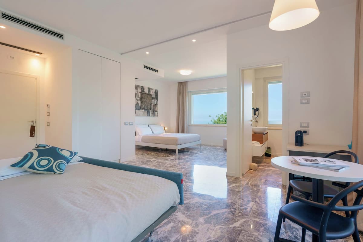 A modern hotel room with a bed, a dining area, and a view of the outdoors.
