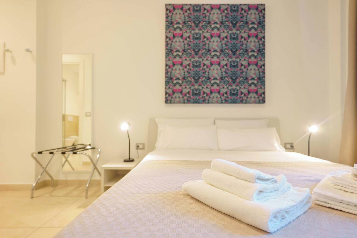 A hotel room with a bed, a wall-mounted artwork, a bedside table with a lamp, and a folded towel on the bed.