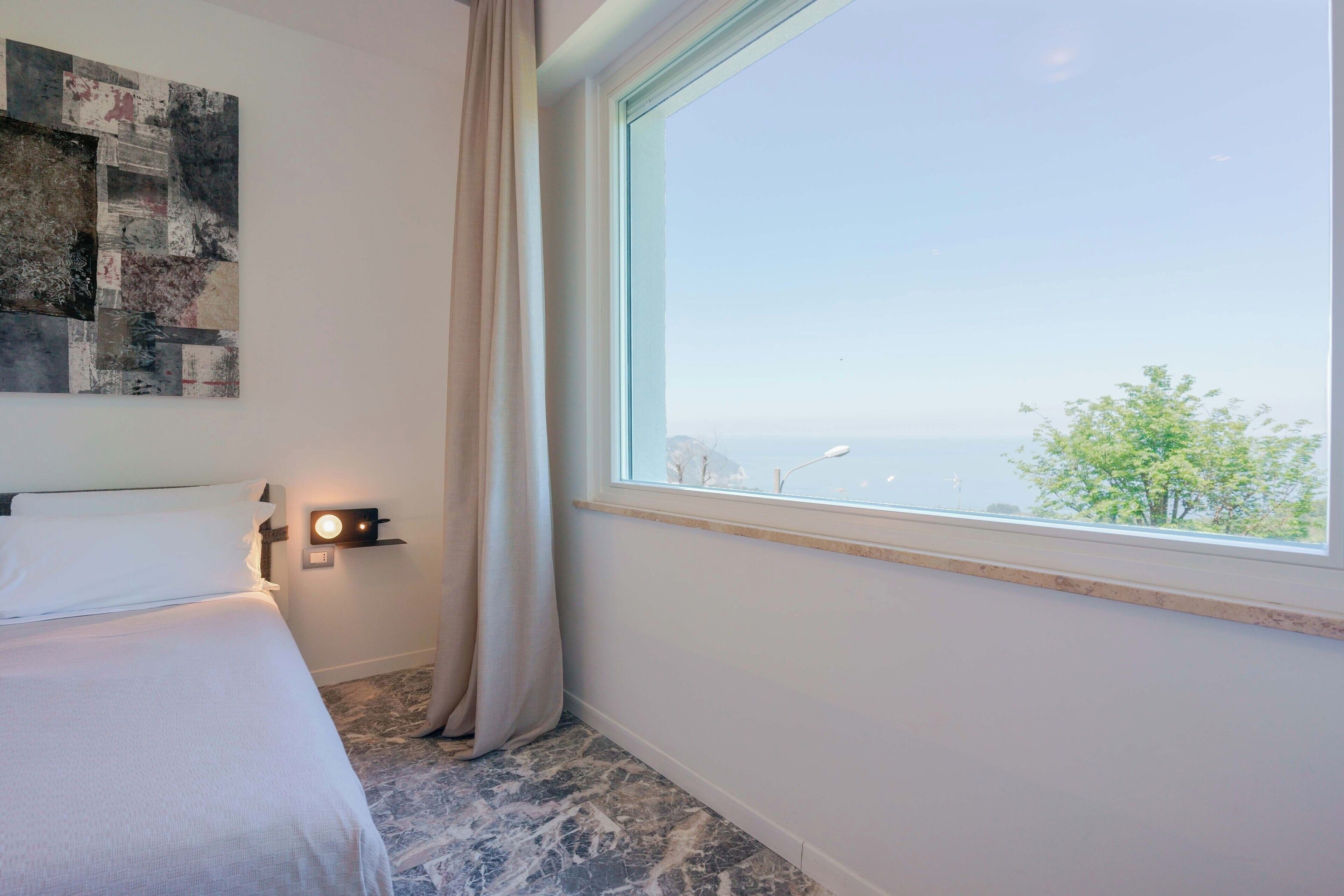 Ein Hotelzimmer mit Bett, Nachttisch mit Lampe, Fenster mit Blick und Wand mit Kunstwerk.