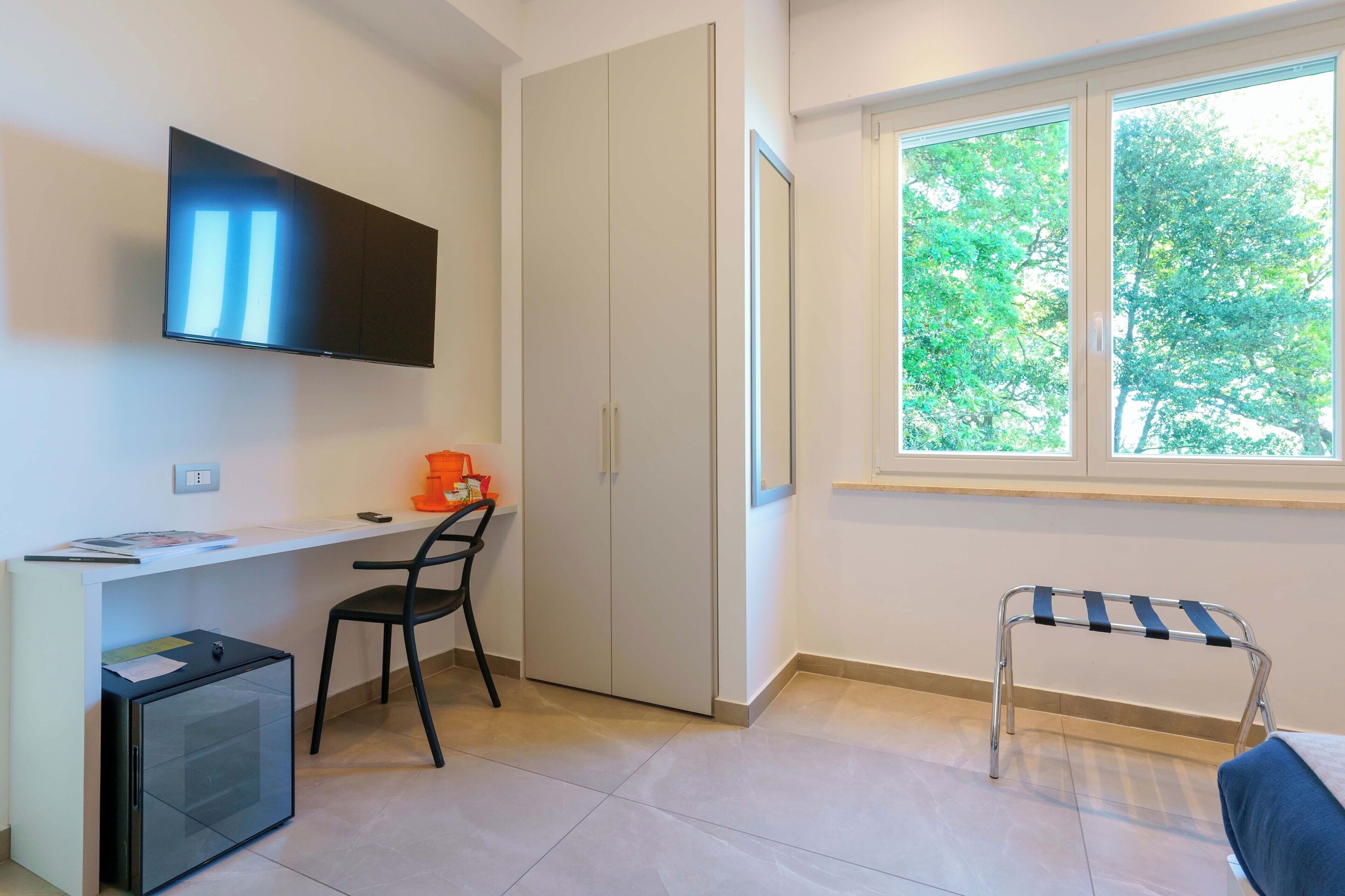 Habitación con televisión montada en la pared, un escritorio con silla, un refrigerador pequeño, armario y ventana con vista a árboles.
