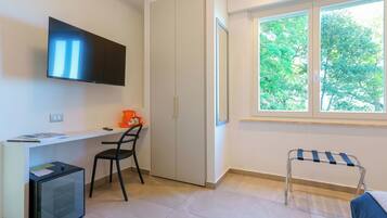 Habitación con televisión montada en la pared, un escritorio con silla, un refrigerador pequeño, armario y ventana con vista a árboles.