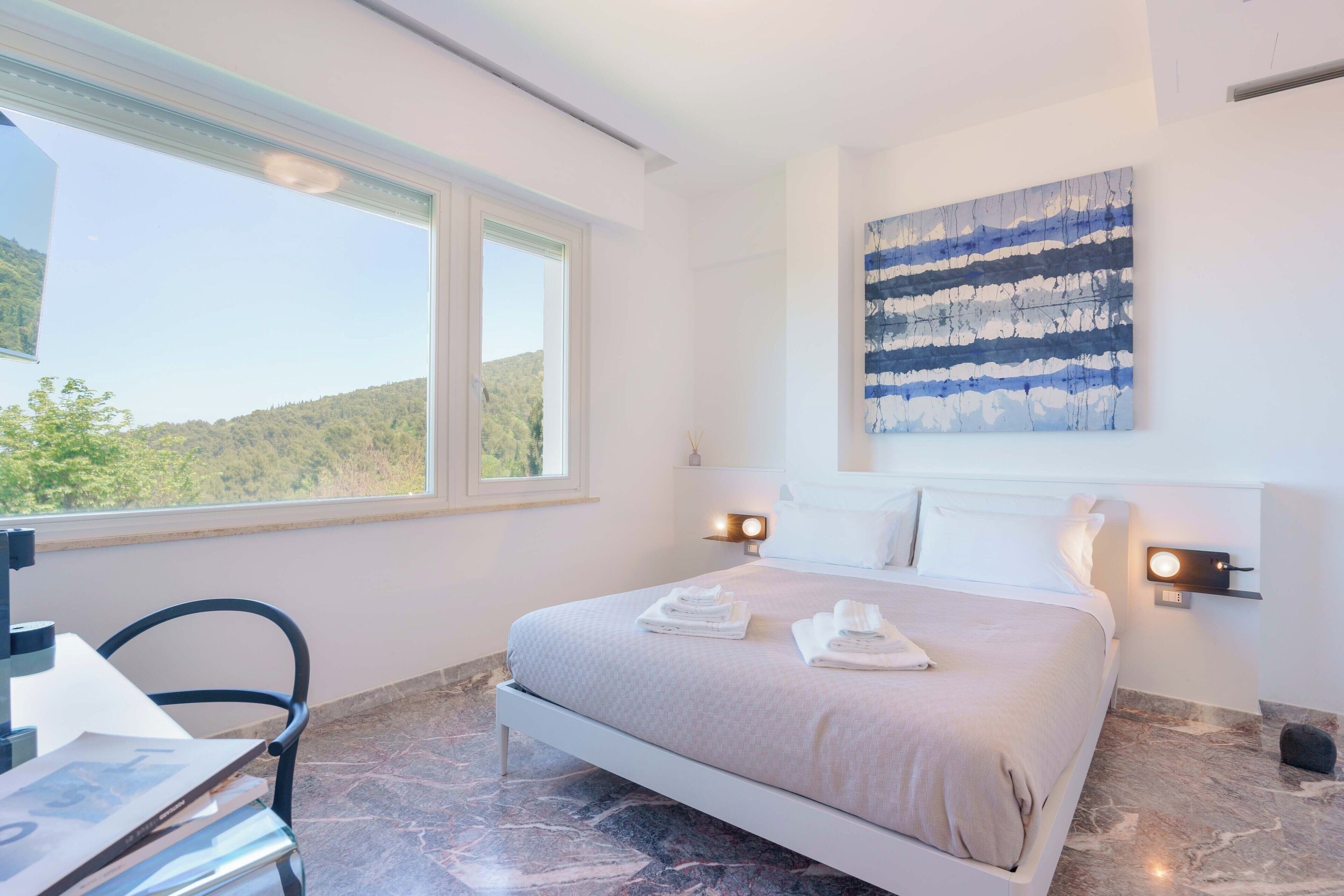 A bedroom with a bed, two bedside lamps, a painting, a window, and a chair.