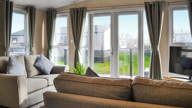 En stue med sofa, fjernsyn og store vinduer med gardiner.