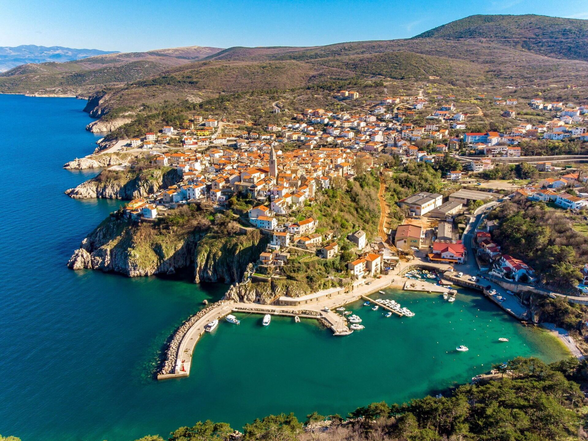 An aerial view of a coastal town with a marina, buildings, and a mountainous backdrop.