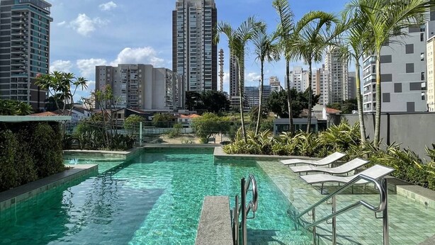 A rooftop pool with clear water, surrounded by palm trees and lounge chairs, overlooking a cityscape with high-rise buildings. - Haus Residence Brooklin (São Paulo)