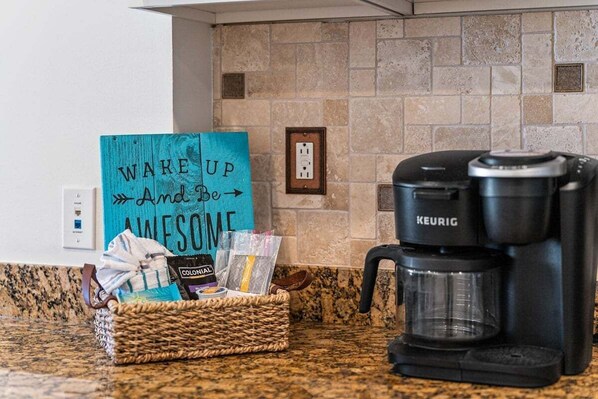 Uma bancada de cozinha com uma cafeteira Keurig, uma cesta de vime com cápsulas de café e um cartão, e um revestimento de pedra.