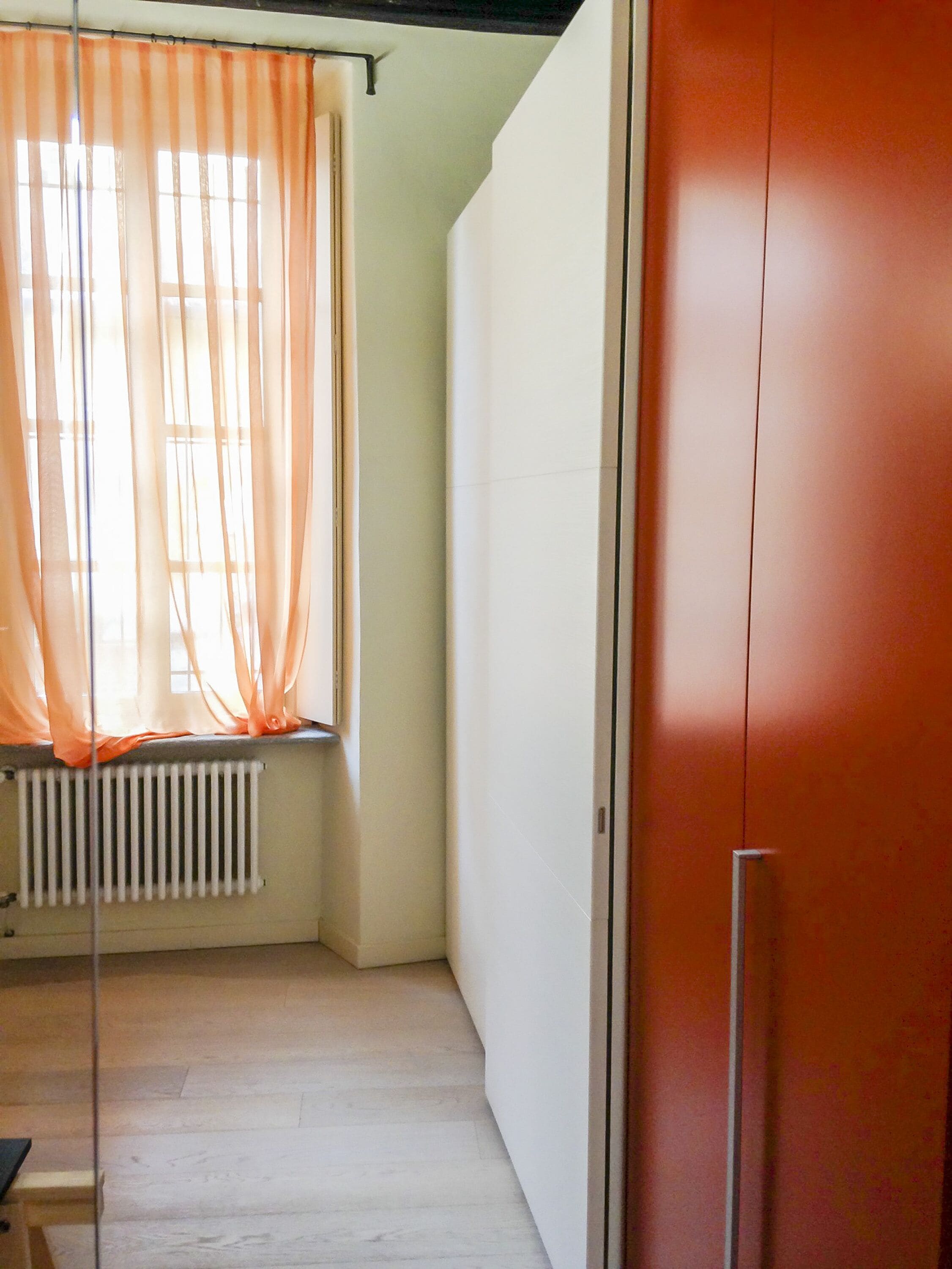 A room with a radiator, a window with sheer curtains, and a red door.