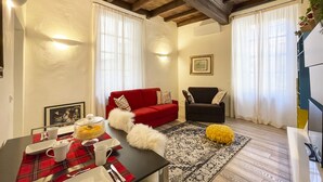 Ein gemütliches Wohnzimmer mit einem roten Sofa, einem schwarzen Sessel und einem Esstisch, der mit einer rot-weißen karierten Tischdecke gedeckt ist.
