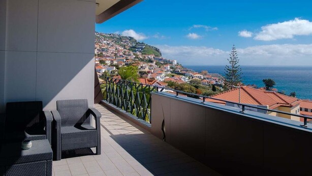 Ein Balkon mit Korbmöbeln, Blick auf eine Küstenstadt und ein klarer Himmel.