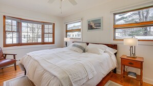 A bedroom with a large bed, wooden headboard, two bedside tables with lamps, a ceiling fan, and large windows with curtains. - 1 Mi to Beach! Family Gloucester Gem w/ Yard (Gloucester)