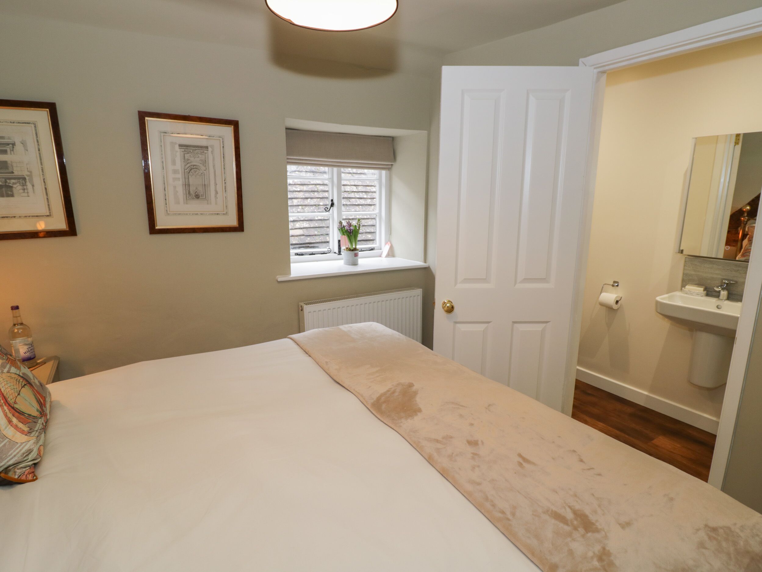 A bedroom with a bed, a window with blinds, a radiator, a door, and a bathroom with a sink and mirror.