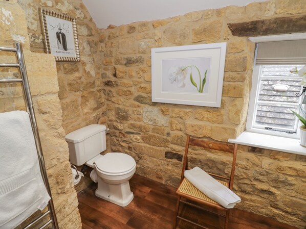 A bathroom with a stone wall, a white toilet, a wooden chair with a towel, and a framed picture of flowers. - The Bindery (Broadway)