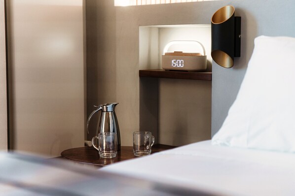 Una habitación de hotel impecable con una mesita de noche, un reloj digital, una cafetera y dos vasos.