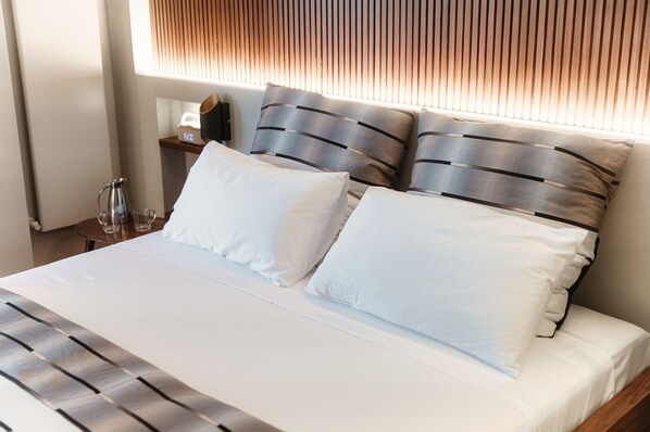 A neatly made bed with white linens and striped pillows, a bedside table with a lamp, and a window with blinds. - Blue Stay (Faggeto Lario)
