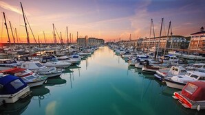 Un port de plaisance où sont amarrés divers bateaux, sur des eaux calmes, et qui est entouré d’immeubles.