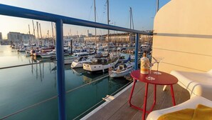 Un balcon donnant sur une marina remplie de bateaux, une table rouge avec une bouteille et des verres, et des chaises longues blanches.