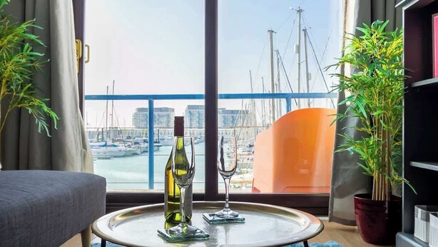A modern living room with a large window overlooking a marina, a round coffee table with a bottle and glass, and a sofa. - Harbour Hideaway - Free Parking (Brighton and Hove)