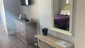Une chambre à coucher avec un lit, une commode, un miroir, une télévision et un bureau avec une poubelle.