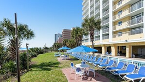 A poolside area with lounge chairs and umbrellas, a grassy lawn, and a building with balconies. - Oceanfront 2Qn Studio. Resort w/Lazy River, Pools (Myrtle Beach)