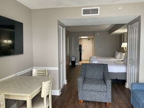 Ein Hotelzimmer mit Esstisch, grauem Sessel, einem Bett mit weißen Leinen und einem an der Wand montierten Fernseher.