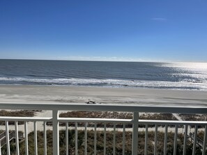 Une vue sur la plage depuis un balcon doté d’une balustrade, avec les vagues de l’océan et un ciel dégagé.
