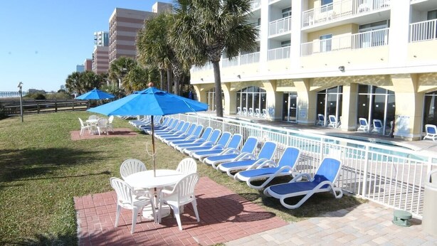 Un espace aménagé au bord de la piscine, avec des chaises longues et des parasols, une terrasse en briques et un bâtiment doté de balcons.