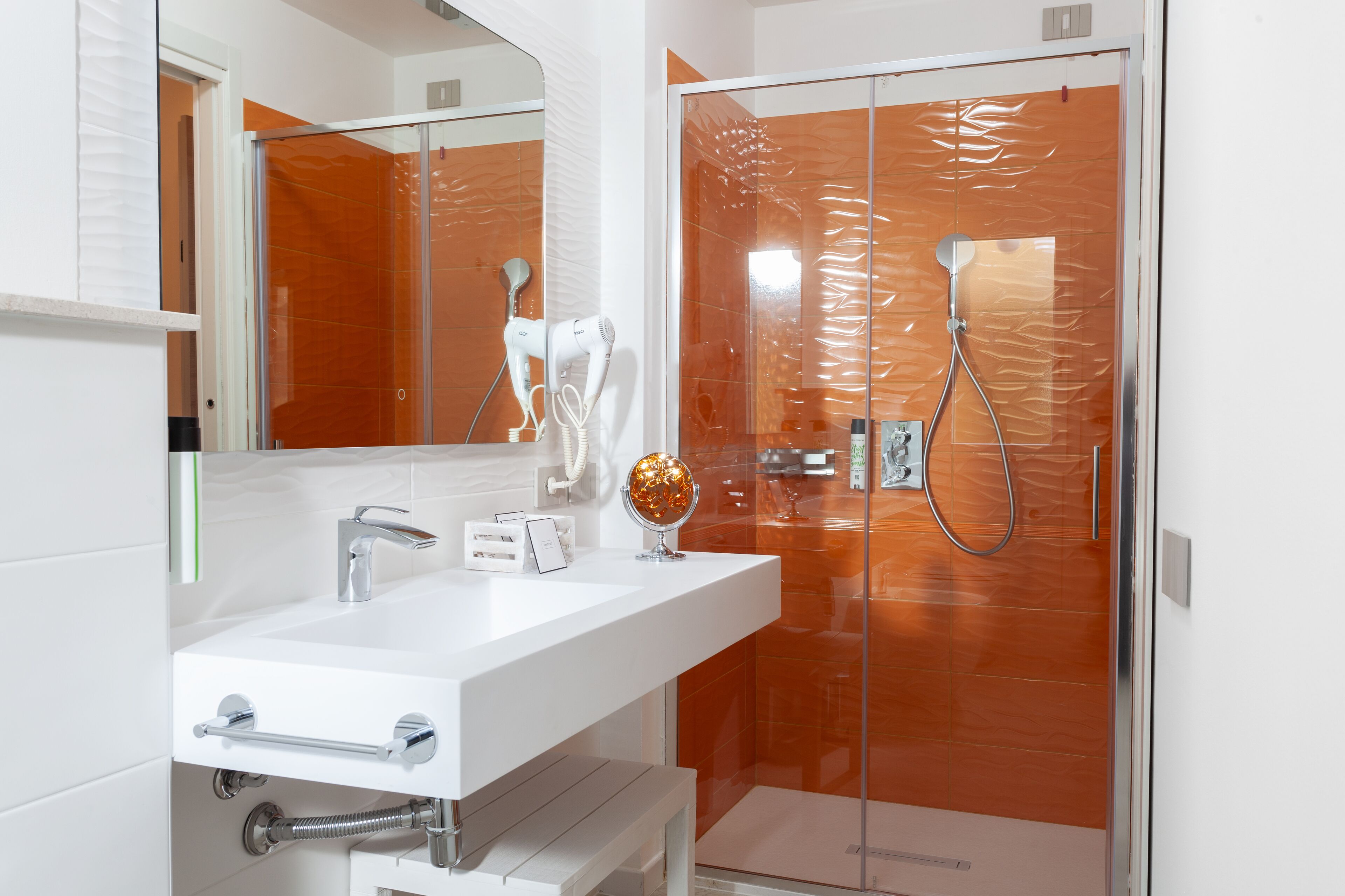 Un baño moderno con un lavamanos blanco, un secador de pelo portátil y una ducha con mampara de vidrio y pared de azulejos naranja.